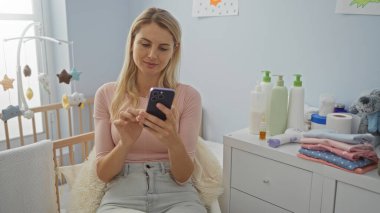 Akıllı telefon kullanan bir kadın yatak odasında beşiği ve bebek ürünleriyle oturuyor, evde huzur verici bir sahne yaratıyor, modern anneliği ön plana çıkarıyor ve ev ortamını rahatlatıyor..