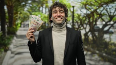 Güneşli bir şehir caddesinde elinde Arjantinli pesolarla gülümseyen bir adam canlı enerji ve yeşillikle dolu bir şehir ortamında pozitif bir mali ruh hali sergiliyor..
