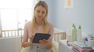Parlak yatak odasında beşiği olan tablet kullanan bir kadın, bebek bakımı malzemeleriyle çevrili, huzurlu bir aile manzarası sergiliyor..