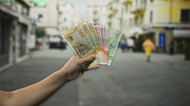 Kalabalık bir şehir caddesinde elinde Norveç kronu banknotları tutan adam, canlı şehir yaşamını ve günışığında keskin detaylarla finansal odağı vurguluyor..