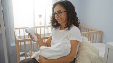 Kadın bebeği yatak odasında tutuyor, arka planda beşik olan termometre kullanıyor, ev ortamında beslenme anını gösteriyor..