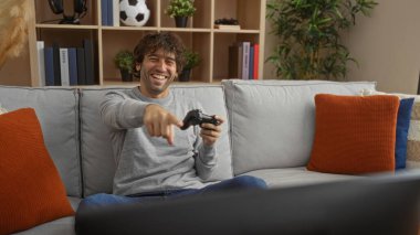Evde video oyunu oynayan genç adam, oturma odasında kanepede oturuyor, kumandayı tutarken gülümsüyor ve işaret ediyor, rahat dekor ve kitaplıklarla çevrili..