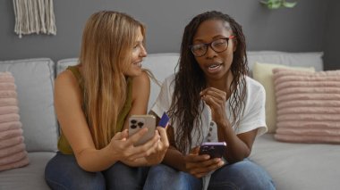 İki kadın rahat bir oturma odasında kanepede oturuyor, ellerinde akıllı telefonlar ve bir kredi kartı, evlerinde yastıklar ve dekorlarla bir anı paylaşıyorlar..