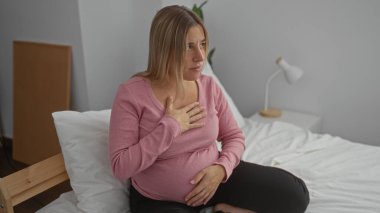 Hamile bir kadın, yatak odasında, üzerinde pembe bluz ve siyah pantolon, göğsünde elle, sakince iç mekanda, hamilelik sırasında rahatsızlık yaşıyor..