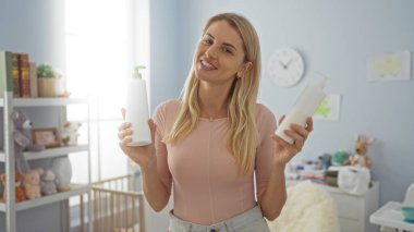 Yatak odasında elinde losyon şişeleriyle gülümseyen bir kadın. Arka planda kreş dekoru olan bir kadın. Bakım ve ev ortamı sunuyor..