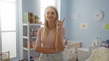 Parlak yatak odasının içini gösteren bir kadın, rahat ev dekorunun ortasında güvenle gülümseyen bir sarışın, odayı doğal ışıkla dolduran davetkar ve neşeli bir atmosfer yaratıyor..