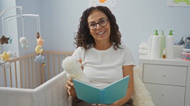 Yatak odasında kitap okuyan bir kadın yanında bebek beşiği olan, sakin bir iç mekan ve neşeli bir atmosfere sahip bir kadın..