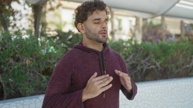 Şehir parkındaki genç İspanyol adam açık havada bilinçlilik egzersizi yapıyor, kapüşonlu giyiyor ve barışçıl bir ifadeyle yeşilliklerin arasında duruyor..