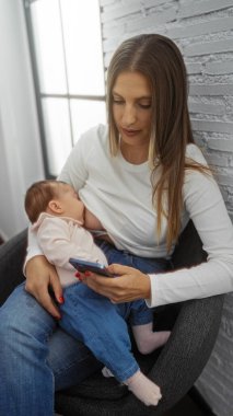 Kadın bebeği emzirirken, ev ortamında rahat bir oturma odası ortamında akıllı telefon kullanıyor, aile bağlarını ve modern ev ortamında birden fazla işi gösteriyor..