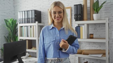 Süslü bir ofiste elinde tabletle gülümseyen bir kadın. Kafkasyalı, günlük mavi gömlekli, arka planda kitap rafı olan bir yetişkin.
