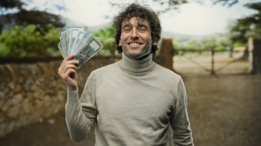 Güneşli bir parkta bangladeş banknotlarını tutarken gülümseyen bir adam doğal bir arka planda neşe ve finansal başarıyı yakalıyor..