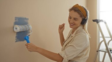 Yeni evin oturma odasında tekerlekli sandalyeyle duvar boyayan, kulaklık takan ve gülümseyen, parlak modern apartmanda ev dekorasyonunun keyfini çıkaran bir kadın..