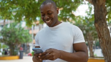 Şehir parkında cep telefonuyla gülümseyen genç bir adam ağaçların arka planında yeşil çevre ve modern yaşam tarzıyla rahat ve mutlu bir anı yakalıyor..