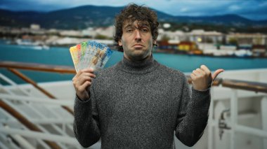 Deniz kıyısındaki bir teknede İspanyol bir adam İsviçre frangı taşıyor, manzaralı bir şekilde çevrelenmiş, kendine güvenen bir ifadeyle yandan işaret ederken..