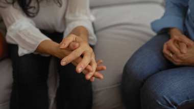 Kadınlar sıcak bir oturma odasında birlikte otururken, elleri destek ve dostluk jesti ile birbirine kenetlenir, samimi bir konuşma ve bağ kurma anını önerirler..