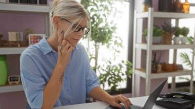 Gözlüklü genç sarışın kadın ev dekorasyon mağazasında dizüstü bilgisayarda çalışıyor. Arka planda bitki ve raflar var.