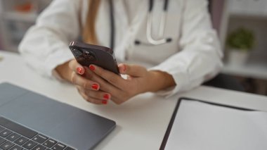 Klinikte cep telefonu kullanan bir kadın steteskobu görünür hale getirerek iletişim teknolojisine odaklanan profesyonel bir sağlık ortamını resmediyor..