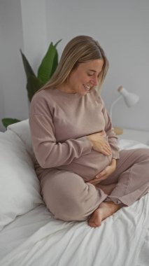 Sarışın hamile bir kadın, yatağın içinde oturmuş karnını nazikçe tutuyor, sıcak bir yatak odasında neşe ve beklenti içinde..