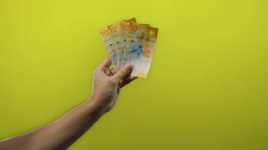 Adamın eli İsviçre frangı banknotlarını canlı sarı bir duvara dayayıp, izole edilmiş para görüntüleriyle finansal temaları vurguluyor..