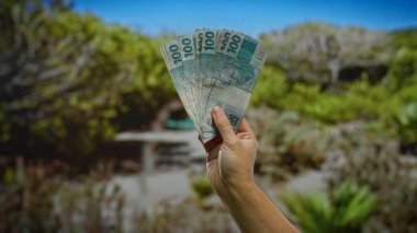 Brezilya 'da güneşli bir şehir parkında el ele tutuşan gerçek banknotlar, Brezilya' da para ve iş kavramını vurguluyor..