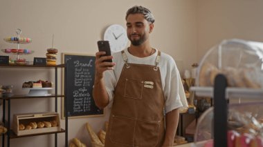 Fırında sakallı genç adam kahverengi önlük giyiyor ve akıllı telefon kullanıyor, raflarda hamur işleri ve ekmek sergileniyor.