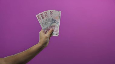 Canlı mor bir arkaplan karşısında Türk lira banknotları tutan adam, izole edilmiş, temiz bir bakış açısıyla finans ve para kavramını resmediyor..