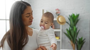 Woman holding baby in cozy living room setting, displaying family love and warmth, with plants and decor creating a peaceful home atmosphere indoors. clipart