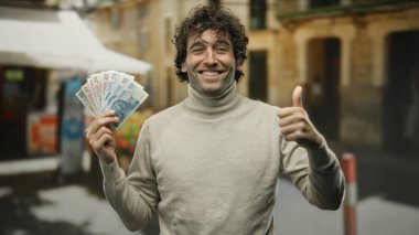 Açık havada gülümseyen bir adam, elinde parlak banknotlar tutarak bir şehir caddesinde baş parmak gösteriyor, coşkulu bir ifadeyi ve kentsel finansal başarıyı vurguluyor..