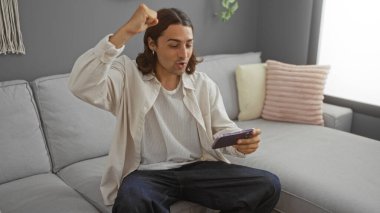 Genç adam modern oturma odasında telefona bakarken, yastıklı rahat bir kanepede otururken kutlama yapıyor..