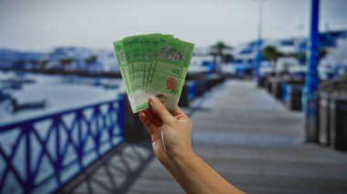 El ele tutuşan Malezya bankası banknotları, deniz kenarı bir arka plan ile, görünürde tekneler ve döviz kuru odaklı bir tatil planı önerir.