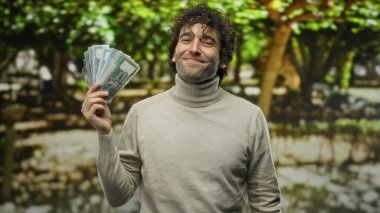 Man smiling holding bangladesh taka outdoors in a sunny park setting displaying happiness and wealth. clipart