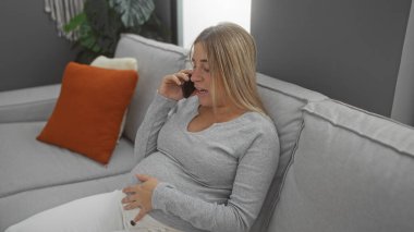 Oturma odasında telefonla konuşan, rahat kanepe ve dekoratif yastıklarla çevrili hamile bir kadın, evde annelik beklentisinin özünü yakalıyor..