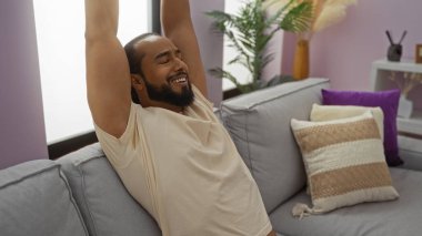 Rahat bir kanepede, modern dekorasyon ve pelüş yastıklı rahat bir evde büyüyen kollarıyla evde dinlenen genç bir adam..