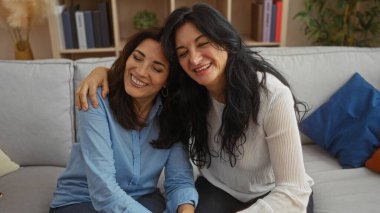 İki orta yaşlı kadın, rahat bir oturma odasında gülümsüyor ve sarılıyorlar. Rahat bir kanepede sevgi ve birlikteliği ifade ediyorlar..