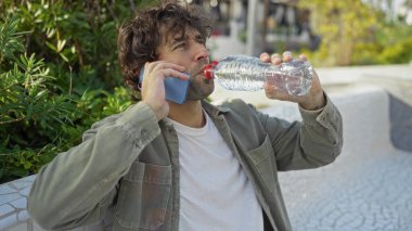 Şehir parkında telefonla konuşurken su içen genç bir adam, çevresi yeşilliklerle çevrili, günlük kıyafetler giyiyor, modern açık hava yaşam tarzını gösteriyor..