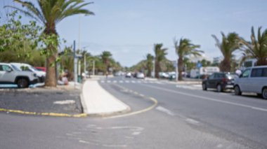 Lanzarote 'deki güneşli bir caddenin bulanık görüntüsü, park etmiş arabaları, palmiyeleri ve dağılmış yayaları olan kanarya adaları..