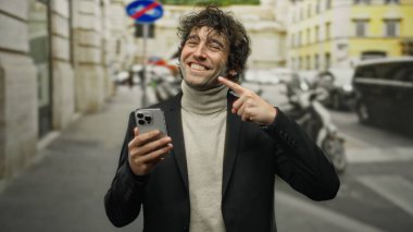 Genç adam bir eliyle dişlerini gösterirken, diğer eliyle akıllı telefon tutarken, şehir merkezinde şık bir ceket giyerek dikilirken gülümsüyor..