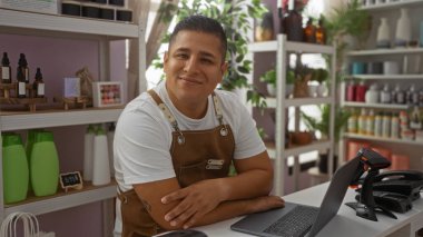 Madalyalı bir mağazada dizüstü bilgisayarı ve çeşitli ürünleri olan çapraz kollu genç bir adam.