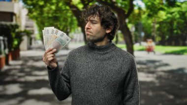 İspanyol asıllı bir adam, şehir dışındaki bir sokakta elinde Czech Koruna banknotları tutuyor. Şehir hayatının güneşli zemininde merak ve tefekkür yansıtıyor..