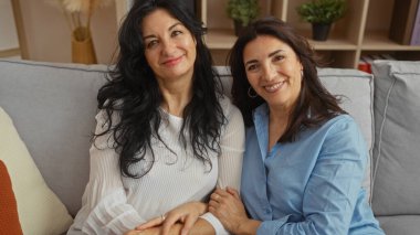 İki olgun kadın, sıcak bir oturma odasında oturup, sıcak bir şekilde kucaklaşıyor ve gülümsüyor, aile sevgisini ve arkadaşlığını bir yuva ortamında sergiliyor, güçlü bir kız kardeş bağını işaret ediyor..