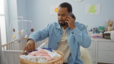 Yatak odasında sakallı bir adam beşiğin yanında çamaşırları sınıflandırıyor ve birden fazla iş ve ebeveynliğin olduğu bir ev sahnesi gösteriyor..