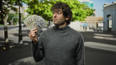 Hispanik bir adam, canlı bir şehir sokak ortamında elinde Amerikan doları banknotları tutuyor. Düşünceli bir ifadeyle, finansal başarısını ve kentsel yaşam tarzını gösteriyor..