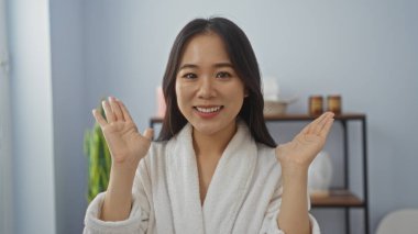 Kadın spa odasında mutlu bir şekilde el sallıyor, beyaz bir bornoz giyiyor ve içini rahatlatıyor, neşesini ve sağlığını vurguluyor.