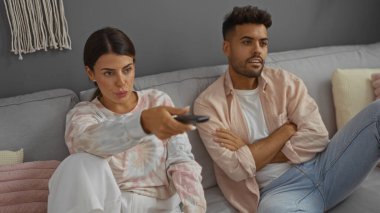 Kadın uzaktan kumandayı gösterirken erkekler modern oturma odasındaki koltukta oturup evde birlikte televizyon izleyen bir çiftin rahatlamış sahnesini tasvir ediyor.
