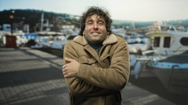 Genç adam limanda, teknelerin arasında üşüyor, ceket giyiyor, yakışıklı, İspanyol asıllı, çekici, deniz kenarı manzaralı, titreyen bir jest yapıyor..