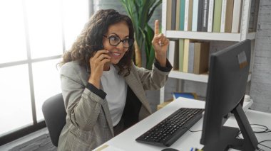 Ofiste telefonla konuşan, gözlüklü ve ceketli bir kadın, bilgisayarlı bir masada otururken, etrafı kitaplıklarla çevrili ve parlak çalışma ortamında bitki yetiştiren birini işaret ediyor..