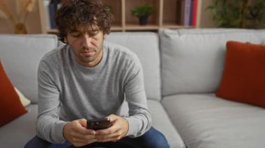 Oturma odasında kanepede oturan genç bir adam, elinde oyun kumandası, yanında akıllı telefon, günlük kıyafetler giymiş, rahat bir ev ortamında video oyunlarına odaklanmış..