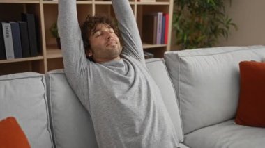 Modern oturma odasındaki kanepede uzanan yakışıklı adam gri süveter ve dekoratif minderlerle şık bir iç mekan ortamında rahatlık ve rahatlık sergiliyor..