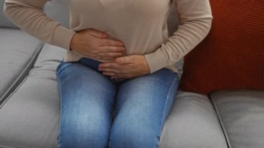 Kanepede oturmuş elleri karnında oturan bir kadın rahat bir oturma odasında kot pantolon ve süveter giyiyor..