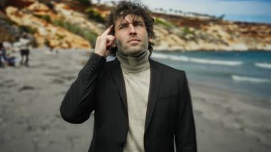 Kıvırcık saçlı genç adam kumsalda bir fikir jesti yapıyor, siyah bir takım elbise giyiyor, düşünceli bir yüz ifadesi sergiliyor ve çarpıcı bir sahil resminde yakışıklı bir görünüm sergiliyor..